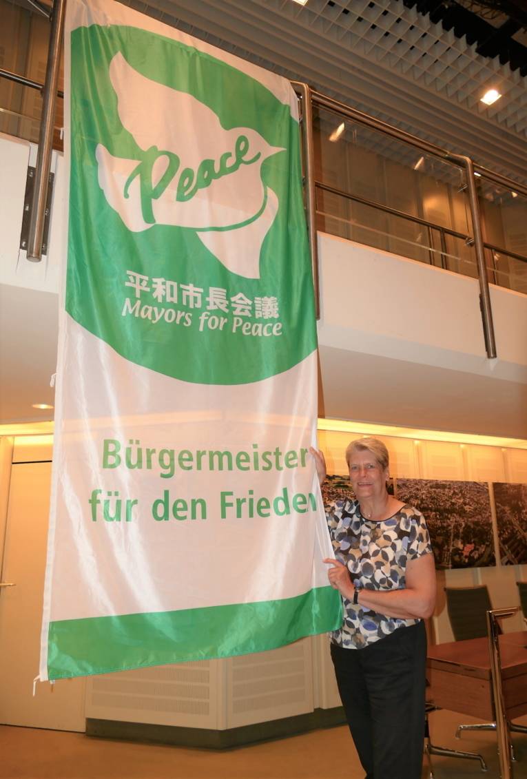 Eine kurzhaarige, blonde Frau hält die Mayors-for-Peace-Flagge in einem Gebäude in Bünde (Nordrhein-Westfalen).